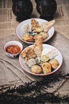 Two bowls filled with various types of fried and steamed snacks, garnished with a slice of lime and served alongside a small dish of red chili sauce. The setup is on a rustic cloth, with an underlying surface of newspaper print and decorative elements in the background.