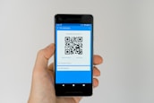 A hand holding a smartphone displaying a QR code on the screen. The interface is primarily blue, indicating a payment or financial app. Above the QR code, there are options related to payment processing.