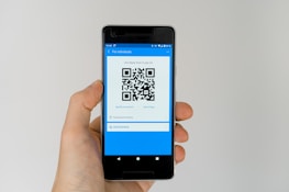 A hand holding a smartphone displaying a QR code on the screen. The interface is primarily blue, indicating a payment or financial app. Above the QR code, there are options related to payment processing.
