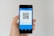 A hand holding a smartphone displaying a QR code on the screen. The interface is primarily blue, indicating a payment or financial app. Above the QR code, there are options related to payment processing.