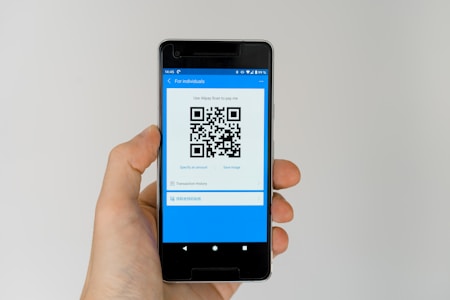 A hand holding a smartphone displaying a QR code on the screen. The interface is primarily blue, indicating a payment or financial app. Above the QR code, there are options related to payment processing.