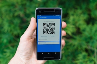 A hand holding a smartphone displaying a QR code. The screen has a blue interface with text indicating it is for a payment application. The background is filled with blurred green foliage.