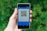 A hand holding a smartphone displaying a QR code. The screen has a blue interface with text indicating it is for a payment application. The background is filled with blurred green foliage.