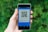 A hand holding a smartphone displaying a QR code. The screen has a blue interface with text indicating it is for a payment application. The background is filled with blurred green foliage.