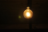 A single incandescent light bulb glows warmly against a dark background, hanging from a fixture, illuminating its surroundings softly.
