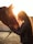 A person with long, curly hair leans in to gently touch noses with a brown horse. The warm glow of the setting sun creates a serene and intimate atmosphere, highlighting the bond between the person and the horse. The person is wearing a black jacket and gloves.