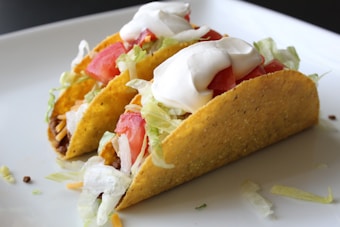 Two hard-shell tacos are filled with seasoned ground meat, shredded lettuce, diced tomatoes, grated cheese, and topped with a dollop of sour cream. They are placed on a clean white plate, highlighting their vibrant colors.