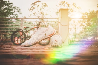 A pair of white Puma sports shoes is placed on a rustic wooden surface. In the background, there is a slightly blurred bicycle and a metal fence. Sunlight creates a warm, colorful lens flare effect, adding a vibrant touch to the scene.