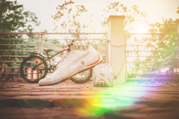 A pair of white Puma sports shoes is placed on a rustic wooden surface. In the background, there is a slightly blurred bicycle and a metal fence. Sunlight creates a warm, colorful lens flare effect, adding a vibrant touch to the scene.