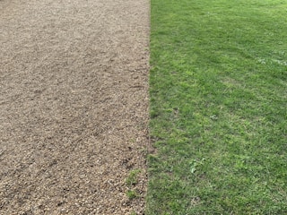 A split landscape with a gravel surface on the left half and a green grass lawn on the right half. The two sections are clearly demarcated and run parallel to each other.