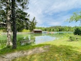 A serene lakeside scene features a wooden cabin by the water's edge, surrounded by lush greenery and tall trees. Small boats are docked at a wooden pier. The sky is partly cloudy, reflecting on the tranquil lake surface. A bench is situated in the foreground overlooking the water.