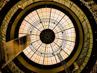 A large, circular glass dome structure with a geometric pattern is viewed from below. The dome is illuminated with a warm, golden light, and a ladder stretches from the center towards the edge. The image includes architectural elements with a futuristic design, featuring interconnected metallic frameworks and a symmetrical, kaleidoscope effect.