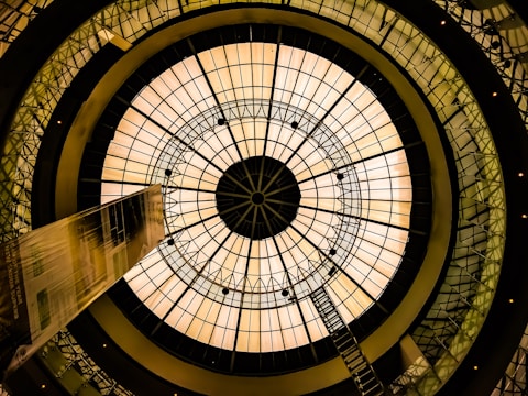 A large, circular glass dome structure with a geometric pattern is viewed from below. The dome is illuminated with a warm, golden light, and a ladder stretches from the center towards the edge. The image includes architectural elements with a futuristic design, featuring interconnected metallic frameworks and a symmetrical, kaleidoscope effect.