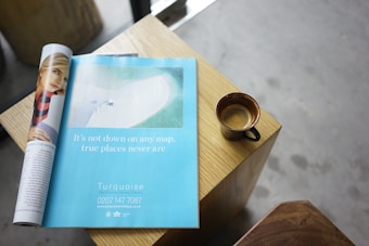 A wooden table holds a rolled-up magazine showing a page with an advertisement. Next to the magazine is a ceramic cup filled with espresso. The ad features a beach scene with text that reads, 'It's not down on any map, true places never are.' The overall setting has a casual, relaxed atmosphere.