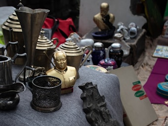 A collection of various antique items displayed on a surface. The assortment includes a golden bust, several metallic pitchers, and intricately designed containers. There are also ceramic teapots, some decorative plates, and a few old boxes with labels and logos, all resting on a gray fabric.
