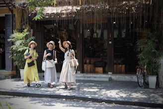 Three women are standing on a sidewalk in front of a shop with wooden doors and potted plants. They are wearing summer dresses and hats, and each is carrying a bag. The shop has a rustic exterior with vines hanging above the entrance.