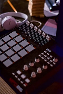 A music production setup featuring a MIDI controller with various knobs and pads, a pair of headphones, a wooden block game, and notebooks. The scene is dimly lit with warm lighting, creating a cozy atmosphere.