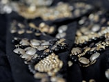 A close-up of intricate beadwork featuring metallic and black beads arranged in an ornate pattern. The details are highlighted by a shallow depth of field, with the focus primarily on the beads in the foreground while the background is blurred.