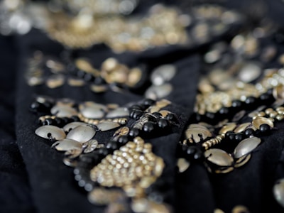 A close-up of intricate beadwork featuring metallic and black beads arranged in an ornate pattern. The details are highlighted by a shallow depth of field, with the focus primarily on the beads in the foreground while the background is blurred.