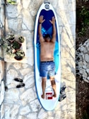 A person lies face down on a paddleboard placed on a stone-tiled surface. The person is wearing denim shorts and has their arms stretched out on the board. Several potted plants are arranged nearby on a low stone wall. A pair of black sandals and a garden tool are positioned on the ground.