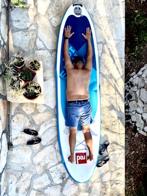 A person lies face down on a paddleboard placed on a stone-tiled surface. The person is wearing denim shorts and has their arms stretched out on the board. Several potted plants are arranged nearby on a low stone wall. A pair of black sandals and a garden tool are positioned on the ground.
