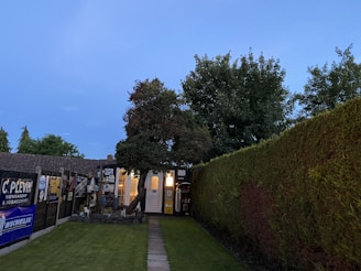 A well-maintained garden path bordered by neatly trimmed hedges leads to a quaint outbuilding with illuminated windows. The area is surrounded by lush green trees and features multiple signs and advertisements on the fence.