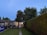 A well-maintained garden path bordered by neatly trimmed hedges leads to a quaint outbuilding with illuminated windows. The area is surrounded by lush green trees and features multiple signs and advertisements on the fence.