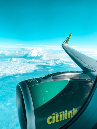A view from an airplane window showcasing the airplane wing and engine with the branding 'Citilink' visible in yellow. The background consists of a vast expanse of clouds and a clear blue sky, giving a sense of travel and elevation.