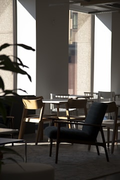 Chairs and tables are arranged in a modern, sunlit room. The area features large windows allowing natural light to cast shadows and highlight the wooden and upholstered furniture. A plant is partially visible, adding a touch of nature to the clean, minimalist design.