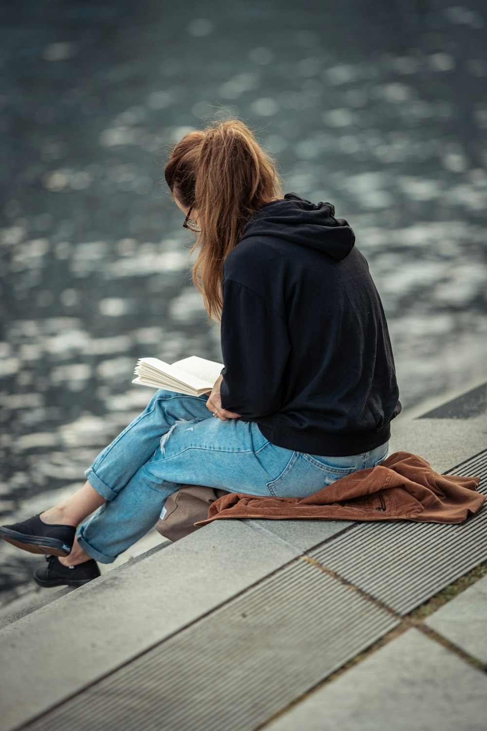 黒いジャケットとブルーデニムジーンズを着た女性が茶色の木製のベンチに座って本を読んでいます