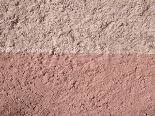 A textured wall with two horizontal sections, the upper part being a light grey color and the lower part in a muted pink. The surface shows a rough, stucco-like texture with visible variations and imperfections.