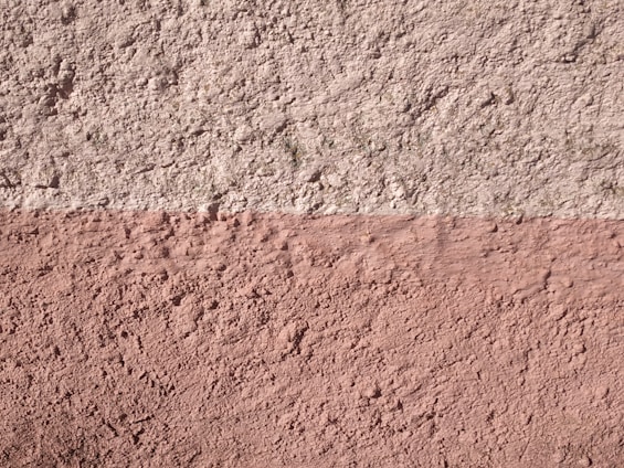 A textured wall with two horizontal sections, the upper part being a light grey color and the lower part in a muted pink. The surface shows a rough, stucco-like texture with visible variations and imperfections.