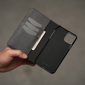 A hand holds a black leather wallet that also serves as a phone case. The wallet is open to reveal a phone slot on one side and a few card slots on the other. There is a dollar bill placed in one of the card slots.