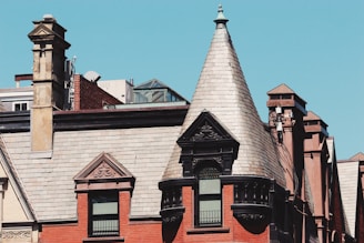 A historic building is characterized by its elaborate brickwork and a prominent conical turret roof. The facade features ornate architectural details with several windows. A stone chimney rises on the side, adding to the vintage aesthetic. The sky is clear and blue, providing a stark contrast to the reddish-brown tones of the building.