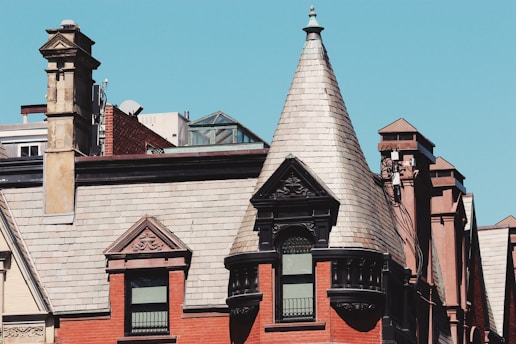 A historic building is characterized by its elaborate brickwork and a prominent conical turret roof. The facade features ornate architectural details with several windows. A stone chimney rises on the side, adding to the vintage aesthetic. The sky is clear and blue, providing a stark contrast to the reddish-brown tones of the building.
