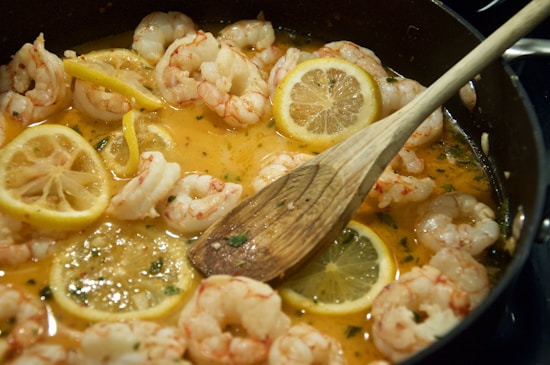 A pan filled with cooked shrimp and lemon slices in a sauce, stirred with a wooden spoon. The sauce has a rich, yellow-orange hue and appears to be seasoned with herbs.