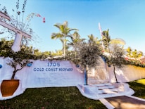 A white stucco wall adorned with the address '1705 Gold Coast Highway' and a sign indicating a private residence. The scene includes lush palm trees, vibrant green grass, and a clear sky. Red flowers and a large potted plant enhance the tropical atmosphere.