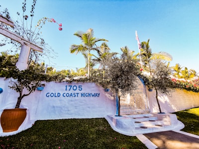 A white stucco wall adorned with the address '1705 Gold Coast Highway' and a sign indicating a private residence. The scene includes lush palm trees, vibrant green grass, and a clear sky. Red flowers and a large potted plant enhance the tropical atmosphere.