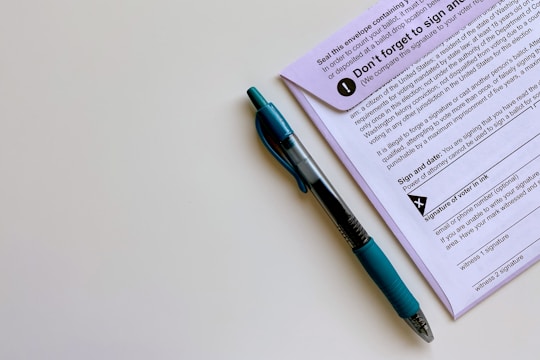 A black and blue pen placed alongside a purple document that appears to contain instructions and a section for signatures. The pen is a retractable ballpoint style with a visible ink chamber and a textured grip. The document includes bold text, bullet points, and a signature area marked with an X.