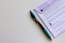 A black and blue pen placed alongside a purple document that appears to contain instructions and a section for signatures. The pen is a retractable ballpoint style with a visible ink chamber and a textured grip. The document includes bold text, bullet points, and a signature area marked with an X.