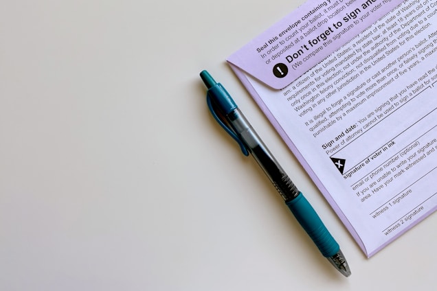 A black and blue pen placed alongside a purple document that appears to contain instructions and a section for signatures. The pen is a retractable ballpoint style with a visible ink chamber and a textured grip. The document includes bold text, bullet points, and a signature area marked with an X.