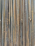 A collection of vertically arranged steel rebar rods displaying a range of rusted and weathered textures. The bars are closely packed and exhibit a pattern of parallel lines and grooves.