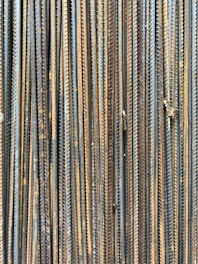A collection of vertically arranged steel rebar rods displaying a range of rusted and weathered textures. The bars are closely packed and exhibit a pattern of parallel lines and grooves.
