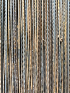 A collection of vertically arranged steel rebar rods displaying a range of rusted and weathered textures. The bars are closely packed and exhibit a pattern of parallel lines and grooves.