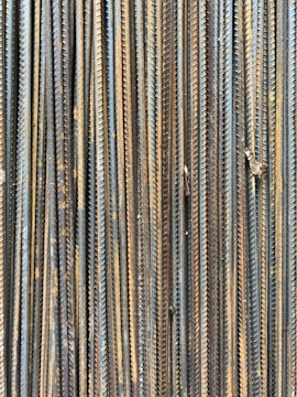 A collection of vertically arranged steel rebar rods displaying a range of rusted and weathered textures. The bars are closely packed and exhibit a pattern of parallel lines and grooves.