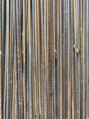 A collection of vertically arranged steel rebar rods displaying a range of rusted and weathered textures. The bars are closely packed and exhibit a pattern of parallel lines and grooves.