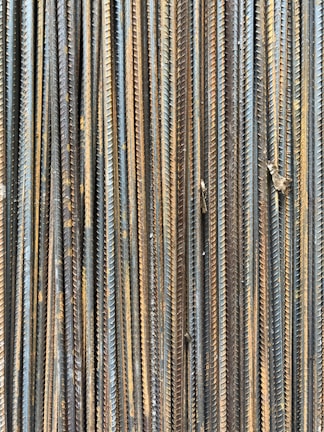 A collection of vertically arranged steel rebar rods displaying a range of rusted and weathered textures. The bars are closely packed and exhibit a pattern of parallel lines and grooves.
