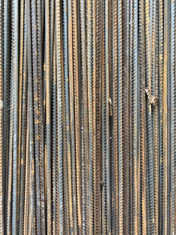 A collection of vertically arranged steel rebar rods displaying a range of rusted and weathered textures. The bars are closely packed and exhibit a pattern of parallel lines and grooves.