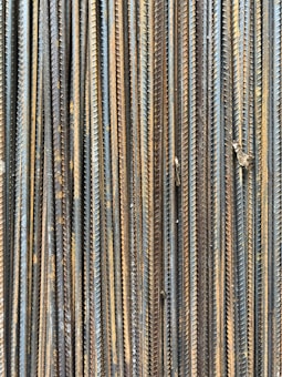 A collection of vertically arranged steel rebar rods displaying a range of rusted and weathered textures. The bars are closely packed and exhibit a pattern of parallel lines and grooves.