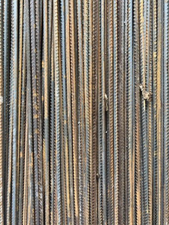 A collection of vertically arranged steel rebar rods displaying a range of rusted and weathered textures. The bars are closely packed and exhibit a pattern of parallel lines and grooves.